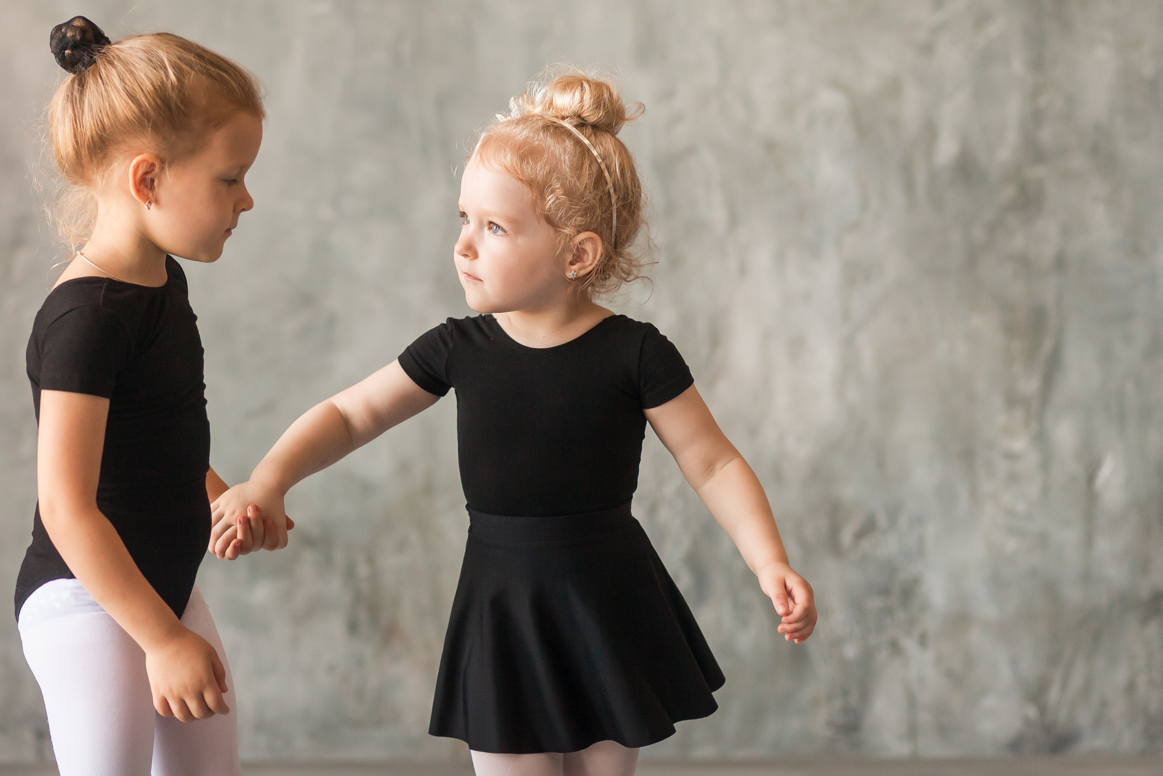 Two small fair-haired girls ballerinas in black packs, white pantyhose, white pointe shoes learn to dance a Russian ballet in a black dance studio