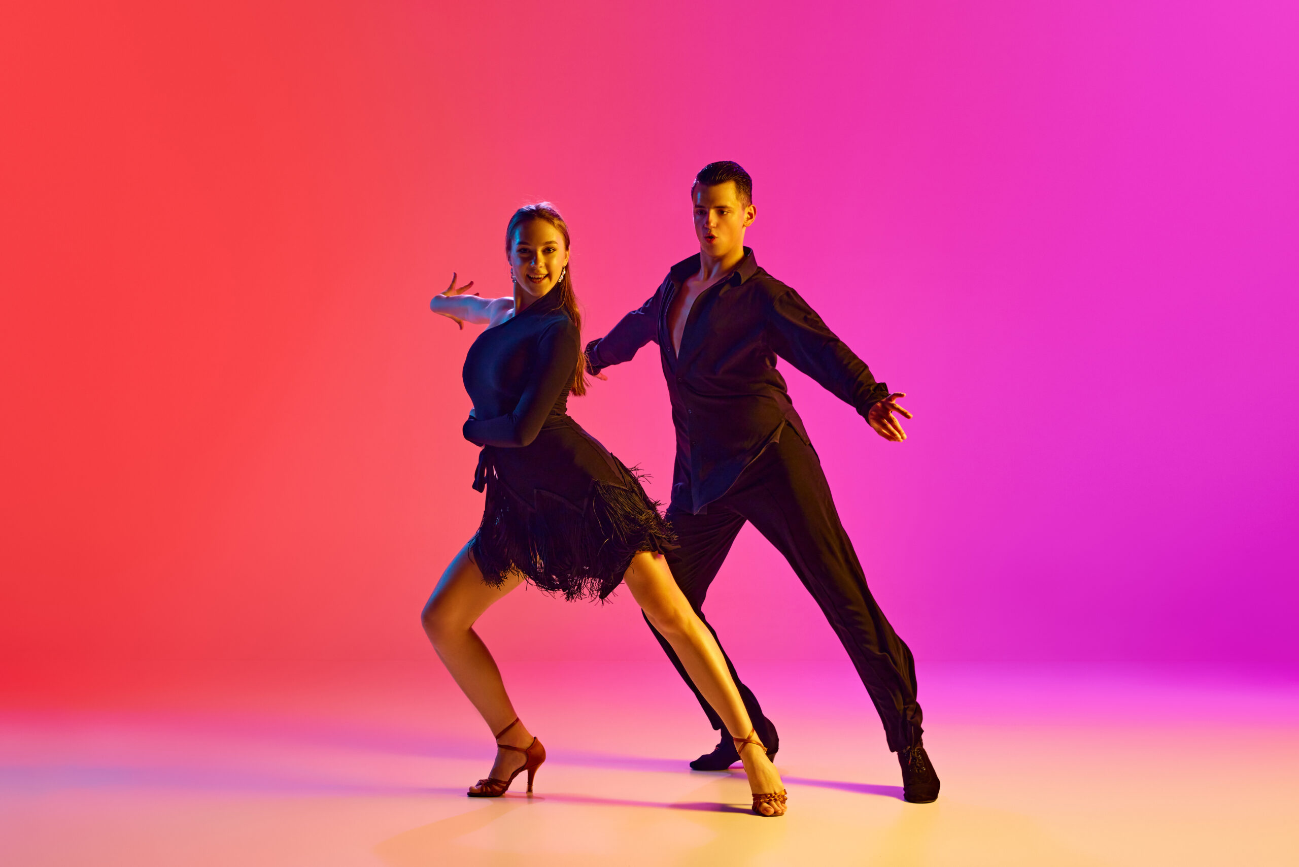 Two young people, man and woman, ballroom dancers display their skills in neon lighting against vibrant gradient background.